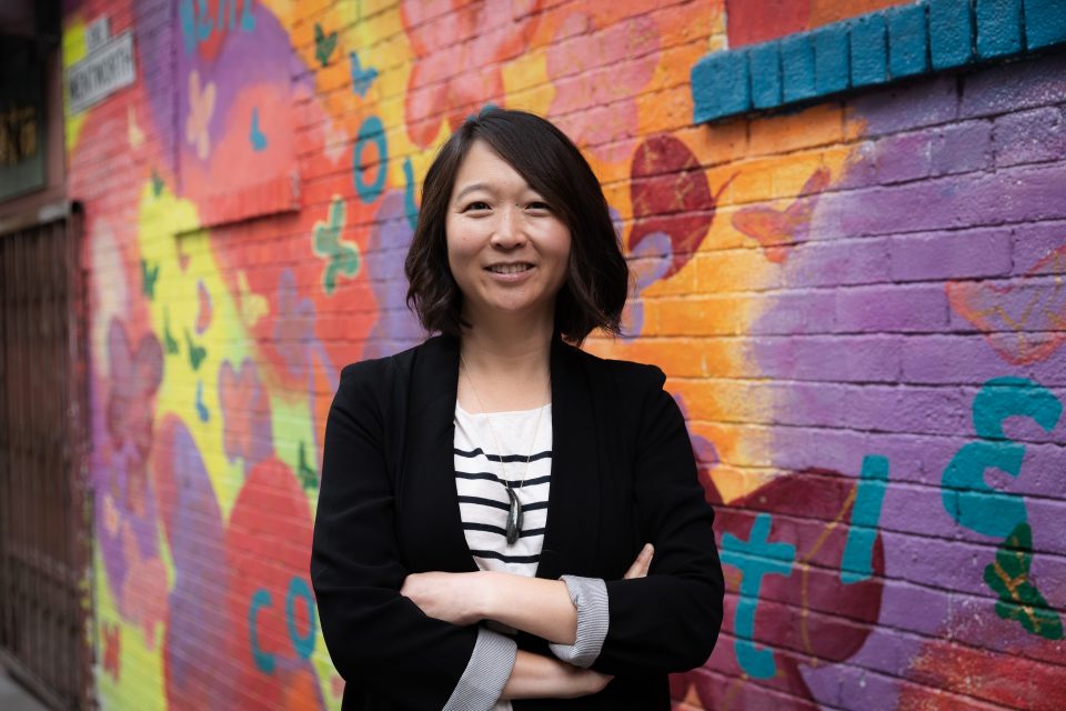 Headshot of Candice Cho: Asian American woman with short, black hair, wearing a black blazer and a striped shirt, smiling at the camera.