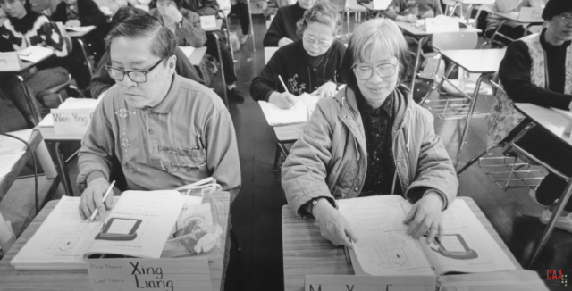 A still of a historical photo from CAA's documentary Block by Block featuring two elders seated on school desks with open workbooks while their classmates, also elders, are writing in their books.