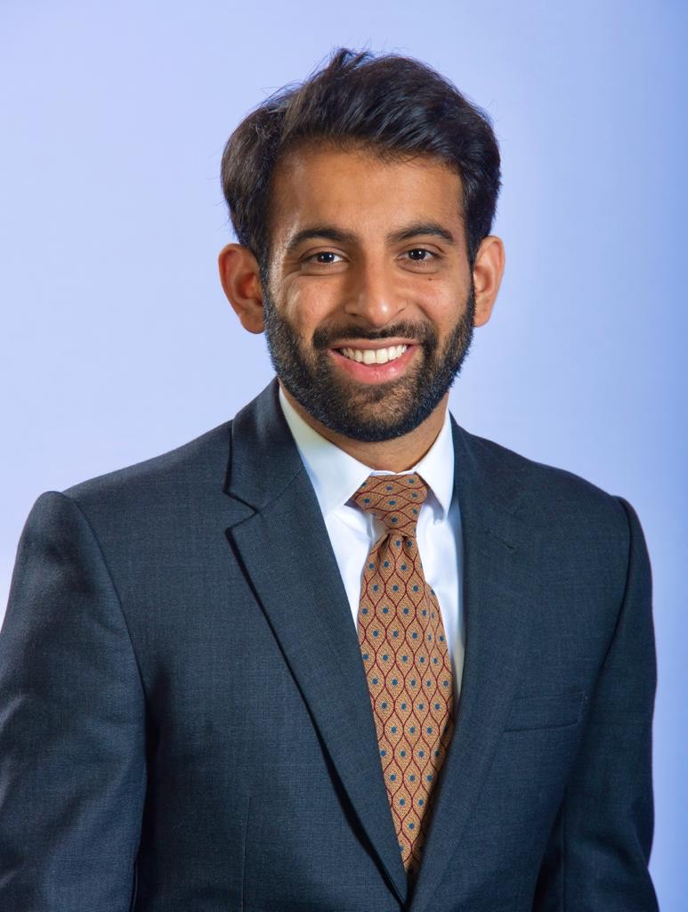 Headshot of Murtaza Headshot, a man with black hair and a black beard wears a suit and tie, smiling.